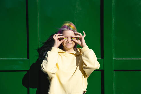 Porträt einer jungen Frau mit gefärbtem Haar und Regenbogen-Bonbons vor einem grünen Container - VPIF02862