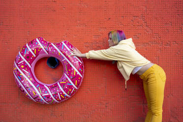 Young woman with dyed hair and floating tyre hanging on red wall - VPIF02874