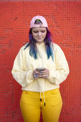 Porträt einer jungen Frau mit gefärbtem Haar, die ein Smartphone vor einer gelben Wand benutzt - VPIF02875