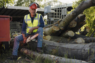 Wrinkled man holding chainsaw while sitting over log in forest - GUSF04354