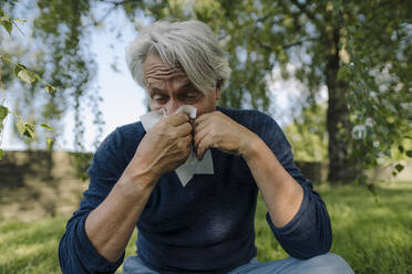Wrinkled man blowing nose while sitting in field - GUSF04390