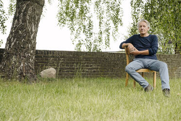 Contemplating man sitting on chair against brick wall in backyard - GUSF04393