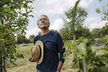 Man holding hat looking away while standing against sky in field - GUSF04423