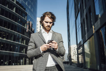 Männlicher Berufstätiger, der ein Mobiltelefon benutzt, während er an einem modernen Bürogebäude in der Innenstadt an einem sonnigen Tag steht - VPIF02899