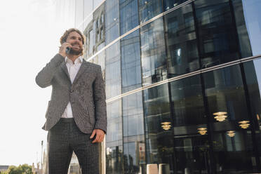 Smiling male entrepreneur looking away while walking on smart phone against office building in financial district - VPIF02912