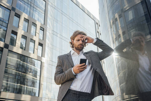 Worried male professional with head in hand using smart phone while leaning by reflection on office building at downtown - VPIF02914