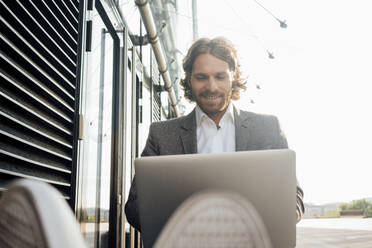 Smiling male entrepreneur using laptop while sitting in downtown on sunny day - VPIF02918