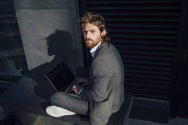 Confident young businessman with laptop sitting on bench at financial district in city - VPIF02919