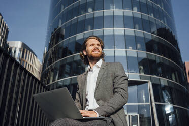 Thoughtful young male entrepreneur sitting with laptop while looking away in financial district - VPIF02921