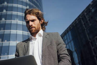 Bearded businessman looking at laptop in financial district on sunny day - VPIF02922