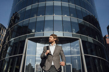 Young male entrepreneur looking away while walking against modern office building at financial district - VPIF02924