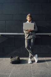Männlicher Unternehmer, der einen Laptop benutzt, während er an einem sonnigen Tag mit gekreuzten Beinen an der Wand steht - VPIF02933