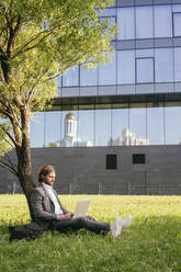 Young male entrepreneur using laptop while sitting against tree on grass at downtown - VPIF02936