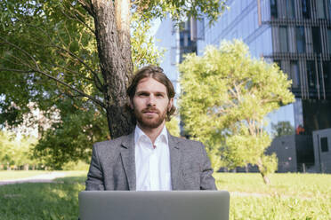 Selbstbewusster, gut aussehender Geschäftsmann, der mit seinem Laptop an einem Baum im Finanzviertel sitzt - VPIF02937
