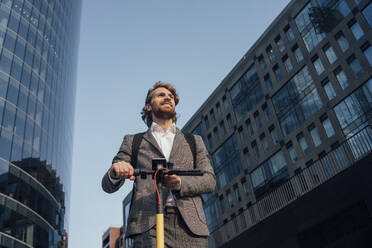 Gutaussehender Geschäftsmann, der wegschaut, während er ein Handy und einen Elektroroller in der Innenstadt hält - VPIF02954