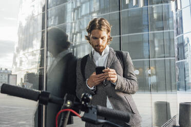 Berufstätiger, der sein Smartphone benutzt, während er sich mit dem Elektroroller im Finanzbezirk an ein Glas lehnt - VPIF02956