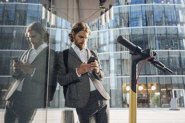 Männlicher Unternehmer, der sein Smartphone benutzt, während er sich mit einem Elektroroller im Finanzbezirk an ein Glas lehnt - VPIF02957