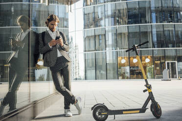 Businessman using mobile phone while leaning on glass with reflection by electric push scooter in financial district - VPIF02958