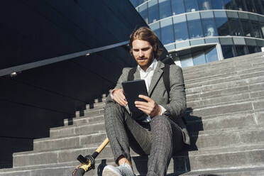 Junger männlicher Unternehmer, der ein digitales Tablet benutzt, während er mit einem Elektroroller auf einer Treppe im Finanzbezirk sitzt - VPIF02968