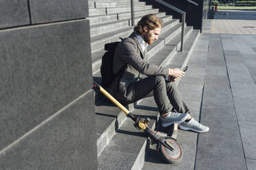 Male commuter using digital tablet while sitting with electric push scooter on steps at downtown - VPIF02969