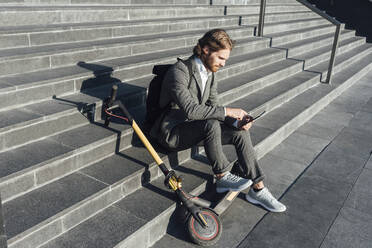 Young businessman with digital tablet and electric push scooter sitting on steps at downtown during sunny day - VPIF02970