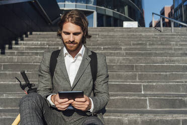 Junger gutaussehender männlicher Profi, der ein digitales Tablet benutzt, während er mit einem Elektroroller auf einer Treppe in der Innenstadt sitzt - VPIF02972