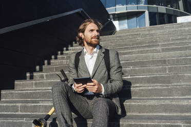 Businessman looking away while sitting with digital tablet and electric push scooter on steps in financial district - VPIF02973