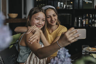Teenager-Freundinnen sitzen am Esstisch, umarmen sich und machen Smartphone-Selfies - MFF06000