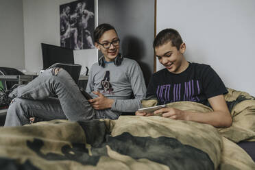 Teenager auf dem Bett sitzend, die sich mit tragbaren Geräten vergnügen - MFF06074