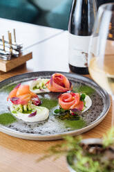 Lachsröllchen mit Zwiebeln und Kräutern, serviert auf einer Metallplatte auf einem Holztisch mit einem Glas und einer Flasche Weißwein - ADSF11750