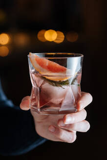 Cropped unerkennbare Person Hand hält Glas mit kalten alkoholischen Cocktail mit Scheibe von Grapefruit und Eiswürfel auf dem Tisch gegen schwarzen Hintergrund platziert - ADSF11758