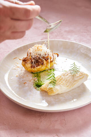 Unrecognizable person with spoon adding cream sauce to yummy fish dish with herb in restaurant stock photo