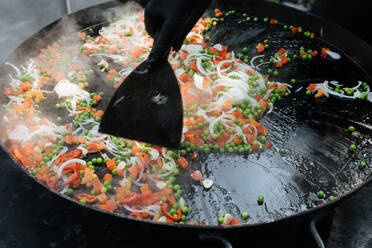 Erntekoch in Handschuh beim Kochen von Erbsen mit Tomaten und Zwiebeln in der Nähe von Garnelen auf einer Metallpfanne auf einem Marktplatz - ADSF11839