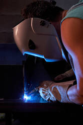 Seitenansicht eines Handwerkers mit Schutzmaske, der einen Overall und Handschuhe trägt und in einer Werkstatt arbeitet und Metallkonstruktionen lötet - ADSF11874