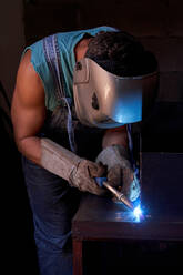 Seitenansicht eines Handwerkers mit Schutzmaske, der einen Overall und Handschuhe trägt und in einer Werkstatt arbeitet und Metallkonstruktionen lötet - ADSF11876