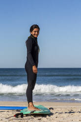 Schöne junge Frau lächelt, während sie auf einem Surfbrett am Strand steht - NGF00599