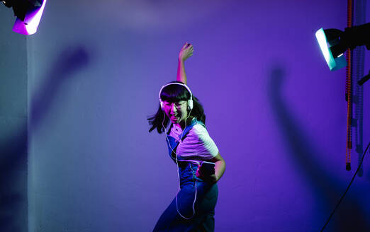 Cheerful woman singing and dancing while listening music through headphones against backdrop - DGOF01246