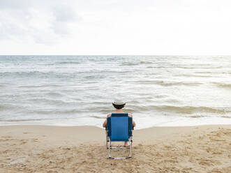 Rear view of man sitting in beach lounger on the beach - JCMF01173