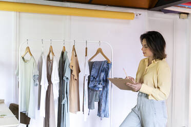 Junge Modedesignerin steht mit einem Klemmbrett und betrachtet Kleidung, die in einem Geschäft an einem Regal hängt - AFVF07029