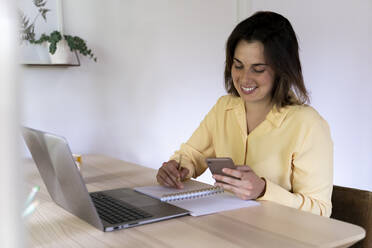 Smiling young businesswoman using mobile phone while sitting with laptop and diary at table - AFVF07049