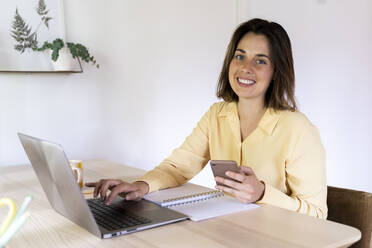 Beautiful female entrepreneur using laptop while sitting with mobile phone and diary at table - AFVF07050