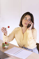 Beautiful young female professional eating salad while talking on mobile phone at home - AFVF07051