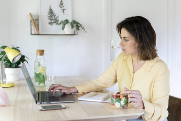 Young businesswoman using laptop while eating healthy salad at table - AFVF07052