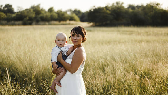 Die Braut trägt einen kleinen Jungen, während sie bei Sonnenuntergang auf einem Feld steht - SAJF00056