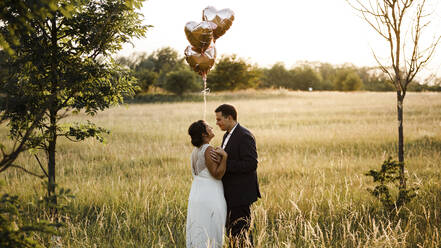 Brautpaar hält Heliumballons in Herzform, während sie in einem Feld bei Sonnenuntergang stehen - SAJF00060