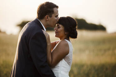 Bridegroom kissing bride on forehead while standing in field during sunset - SAJF00061