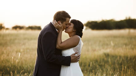 Zärtliches junges Brautpaar, das sich bei Sonnenuntergang auf einem Feld küsst - SAJF00062