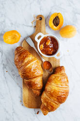 Draufsicht auf ein appetitliches frisches Croissant mit einem Topf hausgemachter Aprikosenmarmelade auf einem hölzernen Schneidebrett in der Nähe von frischem Obst auf einem Marmorhintergrund - ADSF12013