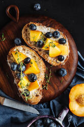 Draufsicht auf appetitliche, gesunde Sandwiches mit hausgemachtem Brot und frischen Aprikosen und Blaubeeren, garniert mit Thymian auf einem Holzbrett auf einem dunklen Marmortisch - ADSF12019