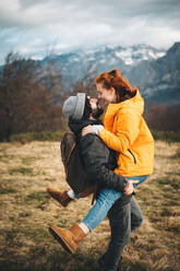 Side view of amorous man with backpack holding smiling woman on hand embracing and having fun in filed with dry grass near by mountains in cloudy day - ADSF12042